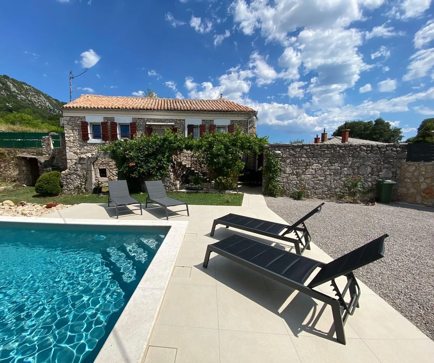 The pool on a clear day, loungers on the deck and the stone house behind